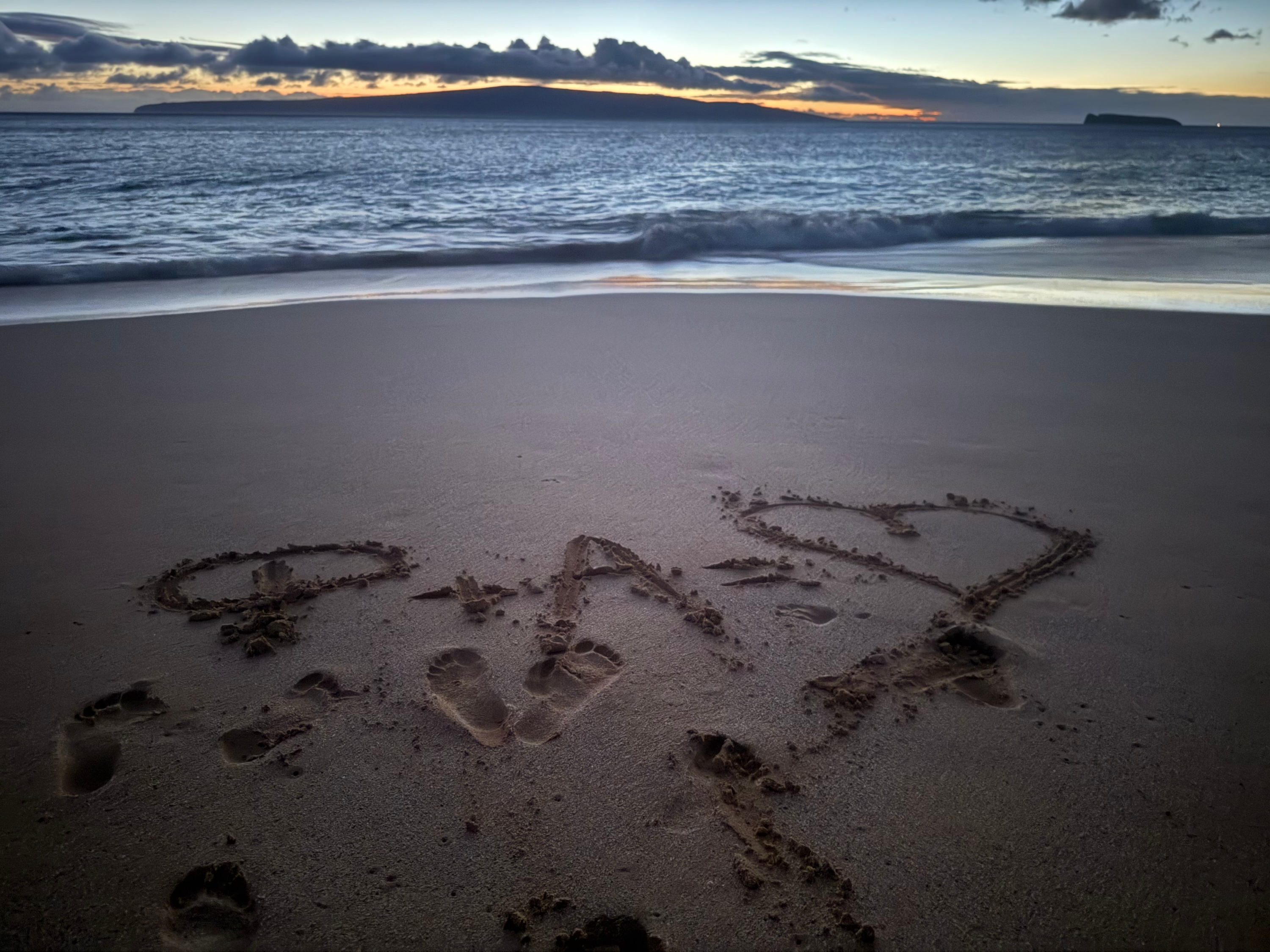 Foivos and Alexandra’s initials and a heart drawn in the sand by the sea at dusk