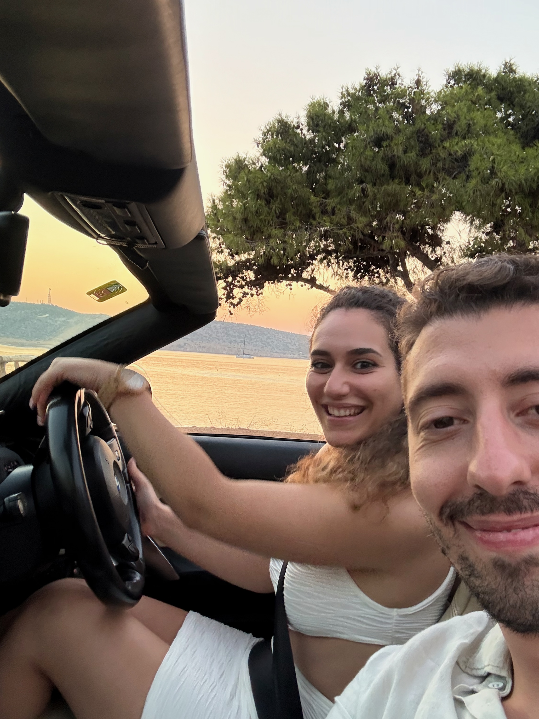 Foivos and Alexandra in a convertible by the sea at sunset
