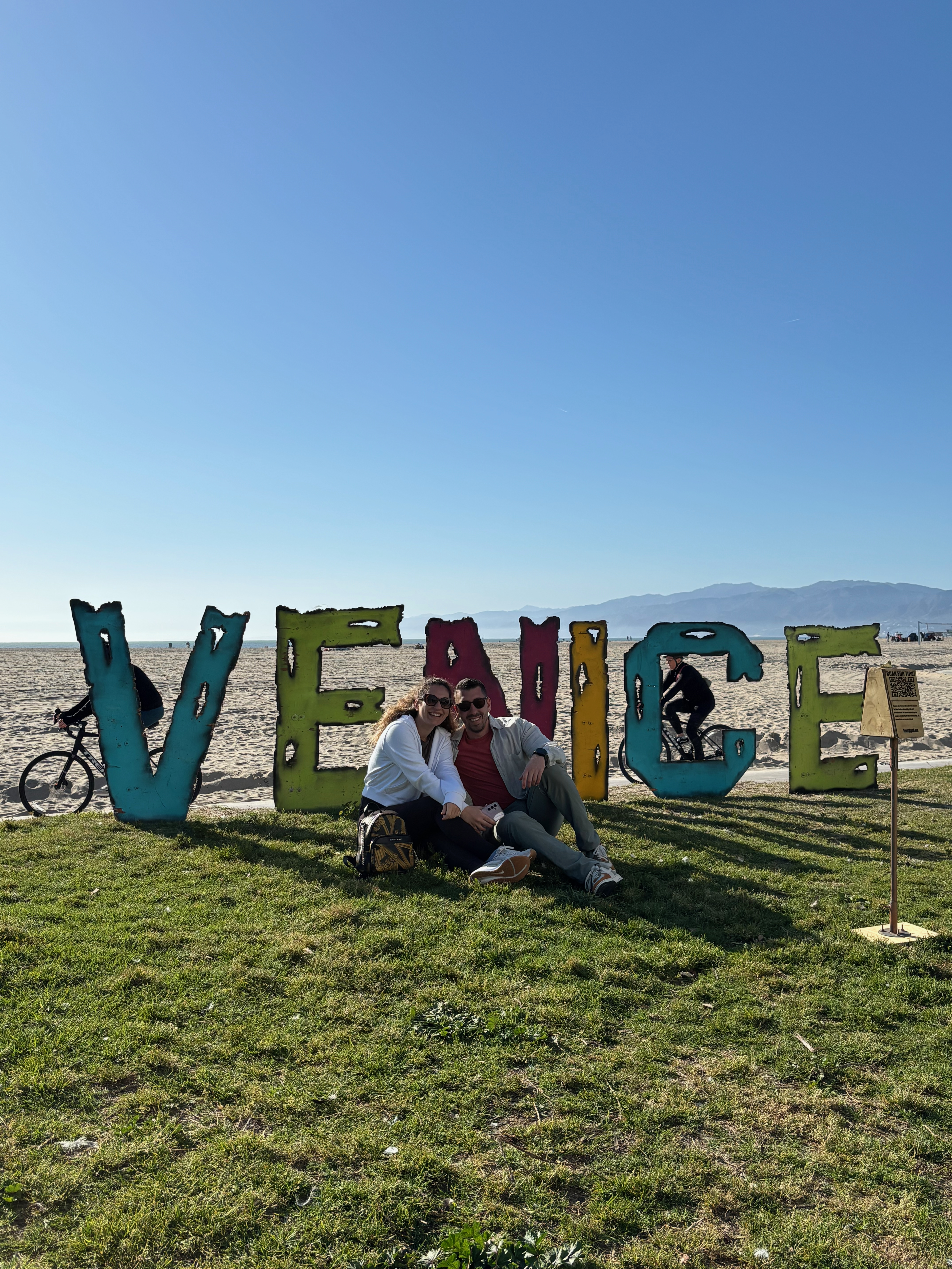 Foivos and Alexandra sitting together in front of the Venice Beach sign